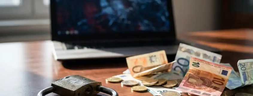 Gebroken hangslot op kantoorbureau naast verspreide eurobiljetten en munten, met beschadigd laptopscherm op de achtergrond.